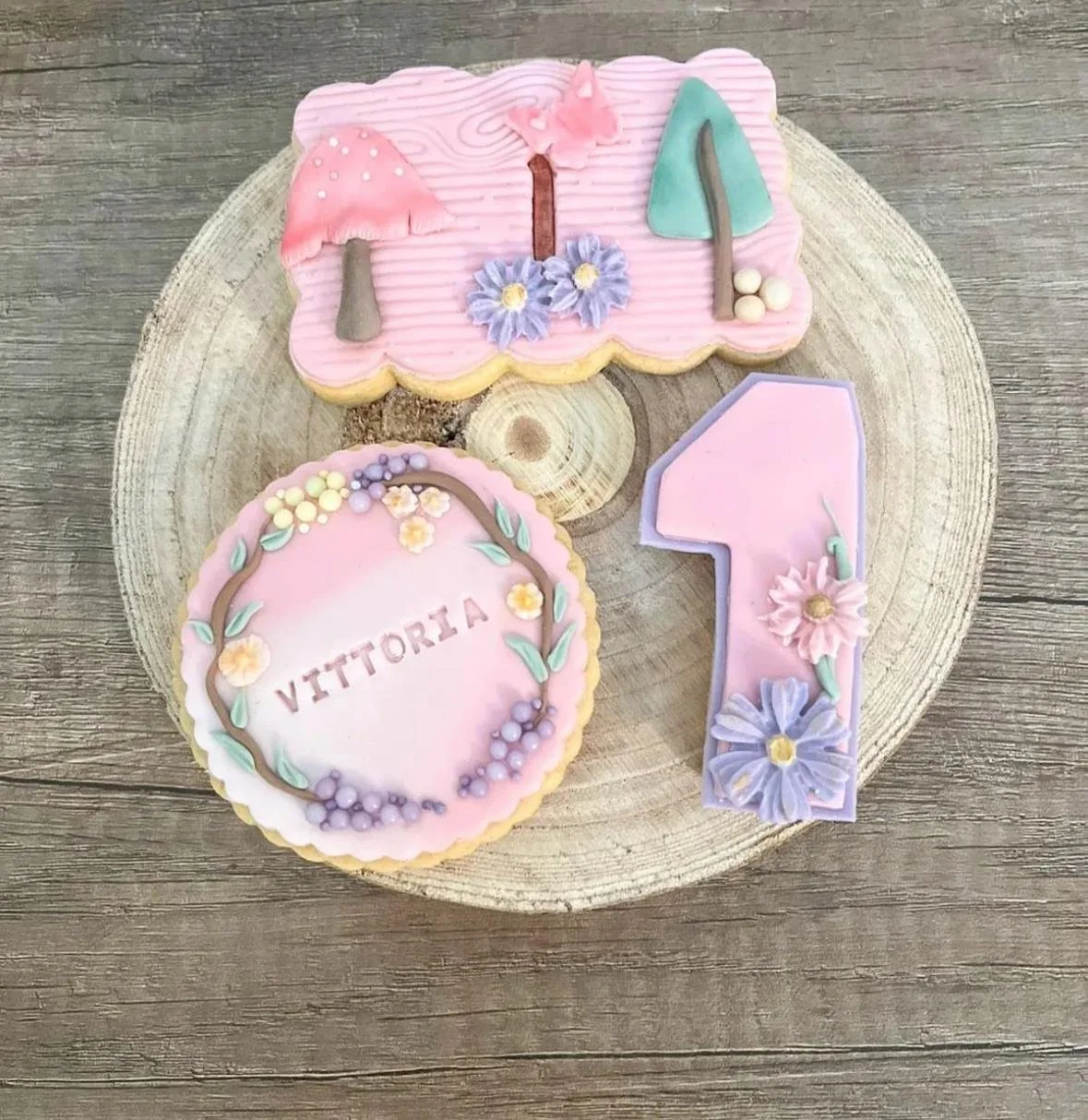 Biscotti decorati con tema festa di primo compleanno, uno con il nome 'Vittoria' circondato da fiori e foglie, un altro con disegni di ombrelli e fiori, e una torta a forma di numero uno decorata con fiori e colori pastel