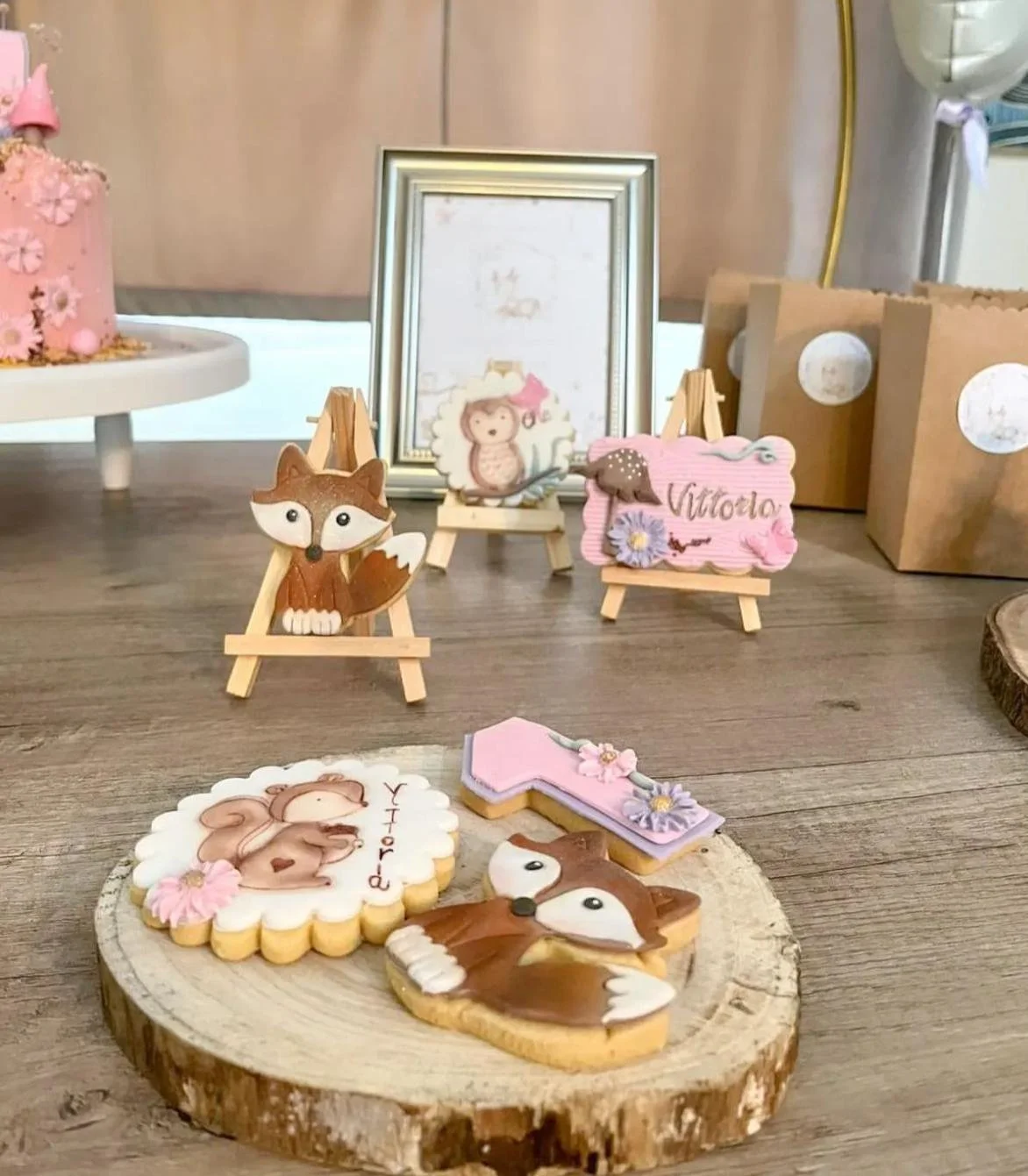 Decorazioni di compleanno con biscotti a forma di scoiattolo e coniglietto, decorazioni con il nome Vittoria, in cornice e alzatina con torta rosa con fiori e coniglietto