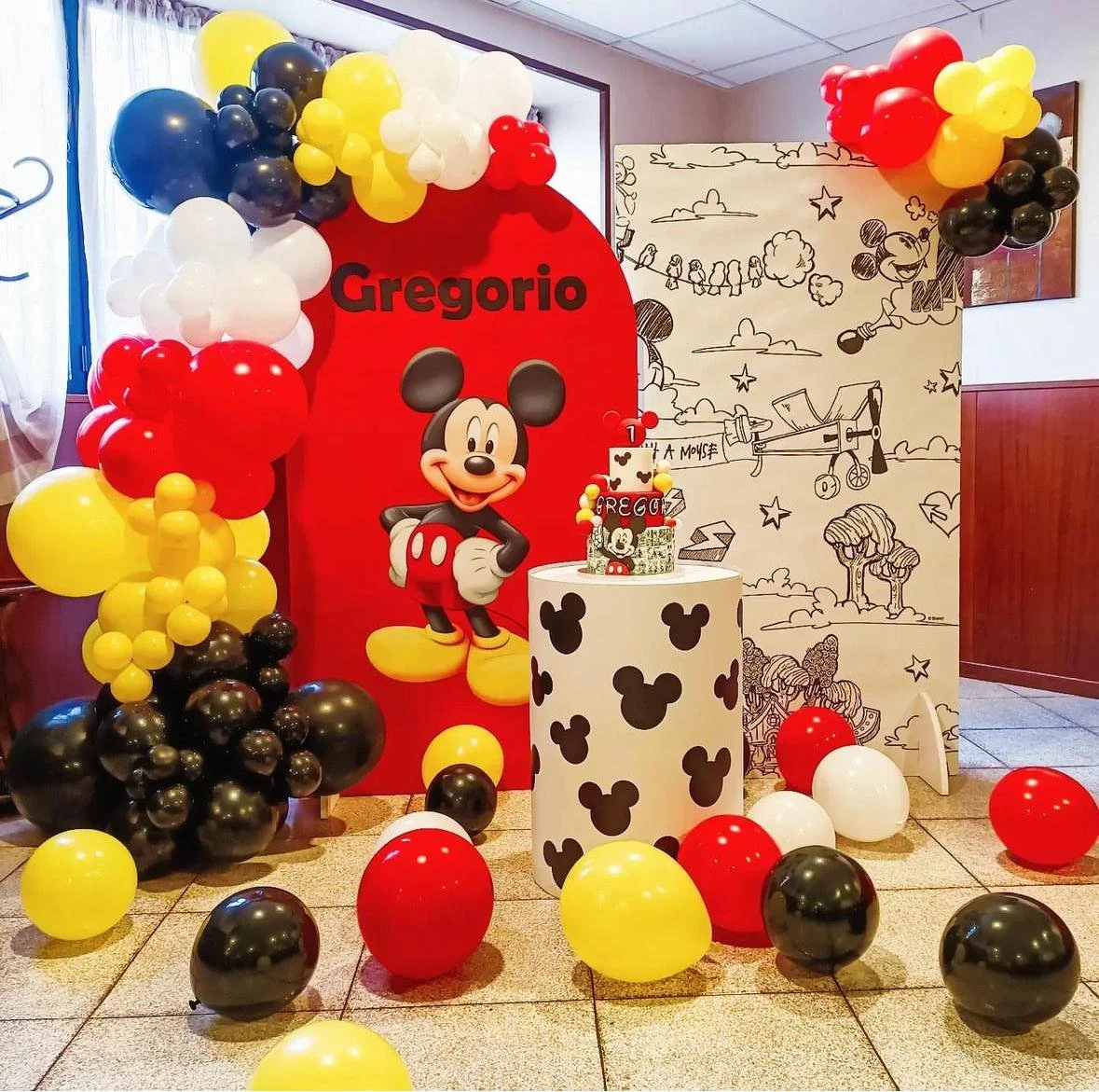 Decorazione di una festa a tema Mickey Mouse con palloncini neri, rossi, gialli e bianco, un grande cartellone con un'illustrazione di Mickey Mouse, una torta decorata con Mickey Mouse e una messa a fuoco di una scritta con il nome Gregorio