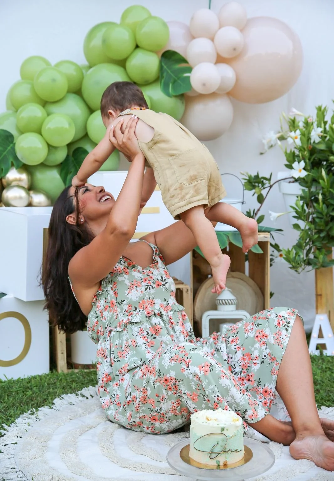 Una donna felice gioca con un bambino in un ambiente decorato con fiori e decorazioni a tema frutta, con un dolce sulla coperta davanti a loro.