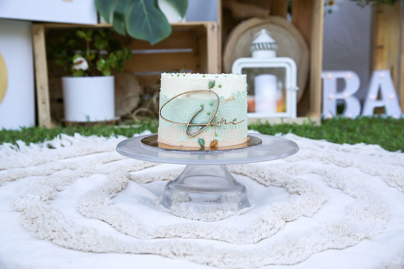 Torta decorata con il nome "One" su un supporto di vetro trasparente, posizionata su un tavolo imbandito con tovaglia bianca decorativa, decorata con piccoli foglie e dettagli dorati, in un ambiente rustico con piante e lettere luminose sullo sfondo.