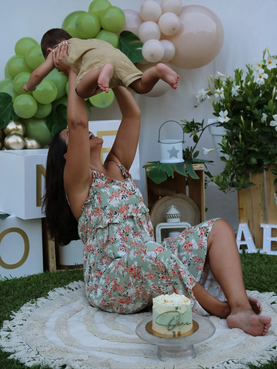 Una donna che solleva un bambino sorridente in aria in un festeggiamento all'aperto con decorazioni di palloncini verdi, bianchI e dorati, e piante verdi, su un prato con un dolce sul pavimento.