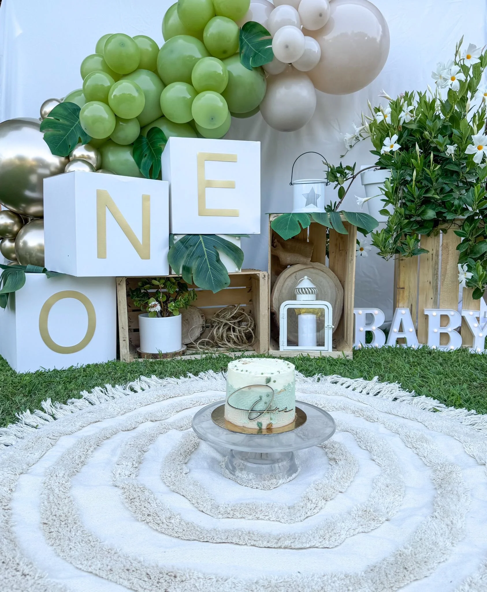 Decorazioni per baby shower con palloncini verdi e bianchi, scatole con lettere che formano 'NOE', fiori bianchi, ciotola con torta decorata con foglie verdi, tappeto bianco e elementi decorativi in stile naturale.